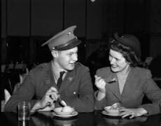 Un couple souriant des années 1940 : un soldat en uniforme militaire et sa petite amie, assise au comptoir d'une fontaine à soda, mangeant une glace (anonyme) - Muzeo.com
